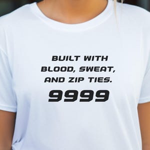 Könnte beinhalten: Weißes T-Shirt mit dem Text "BUILT WITH BLOOD, SWEAT, AND ZIP TIES. 9999" in Schwarz. Das T-Shirt hat kurze Ärmel und scheint aus einem weichen Material zu sein. Das T-Shirt wird von einer Person getragen.