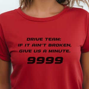 May include: A red t-shirt with the text "DRIVE TEAM: IF IT AIN'T BROKEN, GIVE US A MINUTE. 9999" in black. The shirt has short sleeves and a round neckline.