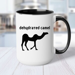 May include: White ceramic coffee mug with a black rim and handle. The mug features a black silhouette of a camel with the text "dehydrated camel" printed on it.