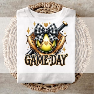 May include: White t-shirt with a gold and black softball design. The softball is in a glove with a bow on top. The text "GAME DAY" is below the softball.