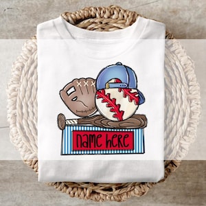 May include: White t-shirt featuring a baseball-themed graphic. The design includes a baseball, a baseball glove, a baseball bat, and a blue baseball cap. The text "NAME HERE" is displayed on a red and blue striped rectangle.