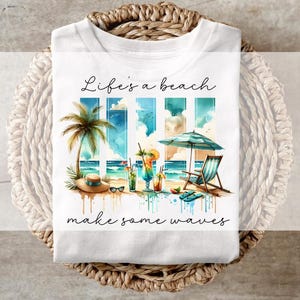 May include: White t-shirt featuring a beach scene with palm trees, cocktails, and a beach chair. The text "Life's a beach make some waves" is written in cursive. The design includes a hat, sunglasses, and a striped umbrella.