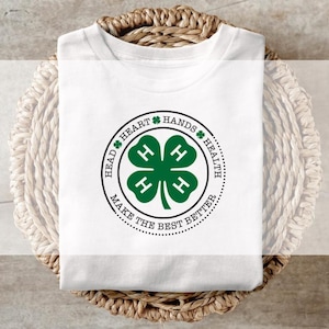 May include: White t-shirt with a green four-leaf clover design. The clover is surrounded by a circle with the words "Head, Heart, Hands, Health" and "Make the Best Better". The shirt is folded and placed on a woven basket.