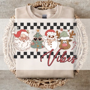 Könnte beinhalten: Beiges Sweatshirt mit Rundhalsausschnitt und Retro-Weihnachtsgrafik. Das Design zeigt den Weihnachtsmann, einen Weihnachtsbaum mit herzförmiger Sonnenbrille, einen Schneemann und ein Rentier, über dem Wort "Vibes" in Rot.