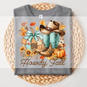 Puede incluir: Camiseta gris con un diseño de temática otoñal. El diseño incluye un sombrero de vaquero, botas turquesas, una calabaza de retazos con un lazo verde azulado, girasoles y el texto "Howdy Fall" en escritura marrón. También se ve una pequeña calabaza naranja.