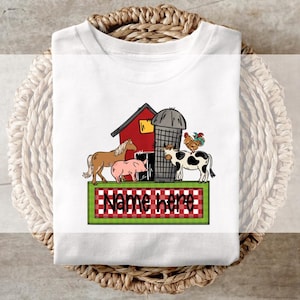 Peut inclure: T-shirt blanc avec une scène de ferme. Il présente une grange rouge, un silo et des animaux de la ferme, dont un cheval brun, un cochon rose et une vache noire et blanche. Le t-shirt porte le texte "Name here" sur une bannière à carreaux verts et rouges.