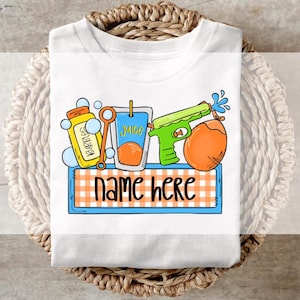 Può includere: Maglietta bianca con una grafica colorata di oggetti divertenti per l'estate, tra cui una scatola di succo, una pistola ad acqua, bolle e una palla da spiaggia. Il testo "name here" è stampato sotto la grafica.