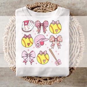 May include: White t-shirt featuring a pink and yellow softball-themed design. The design includes softball bats, balls, gloves, bows, and helmets.