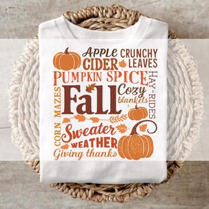 Può includere: Una t-shirt bianca con una stampa grafica in arancione e marrone che raffigura zucche e foglie. Il testo include frasi come Apple Cider, Pumpkin Spice, Fall, Cozy blankets, Corn mazes, Sweater weather e Giving thanks in vari caratteri.