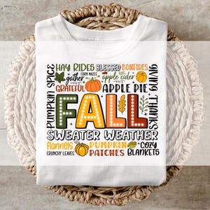Op de afbeelding: Wit T-shirt met een kleurrijk herfstthema. Het ontwerp bevat het woord "FALL" in grote, verlichte letters, omringd door herfstgerelateerde woorden en afbeeldingen zoals pompoenen, bladeren en "sweater weather". Het shirt is opgevouwen in een gevlochten mand.