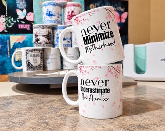 Könnte beinhalten: Zwei weiß gestapelte Keramikbecher mit rosa Kirschblütenmotiven. Der obere Becher trägt den schwarzen Text "never Minimize Motherhood" und der untere Becher zeigt den schwarzen Text "never Underestimate An Auntie".