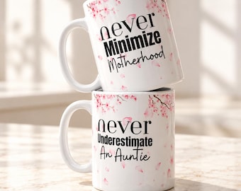 Könnte beinhalten: Zwei weiß gestapelte Keramikbecher, verziert mit rosa Kirschblütenzweigen und Blütenblättern. Der obere Becher trägt den schwarzen Text "never Minimize Motherhood" und der untere "never Underestimate An Auntie".