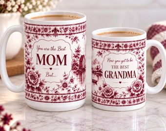 May include: Two white ceramic mugs with maroon floral patterns and landscape illustrations. The larger mug reads, "You are the Best MOM But..." and the smaller mug reads, "Now you get to be THE BEST GRANDMA." Both feature decorative borders and small heart icons.