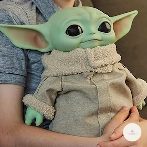 Puede incluir: Una mu&ntilde;eca verde de un personaje con orejas grandes y ojos negros, que lleva un abrigo color canela con cuello y pu&ntilde;os esponjosos. La mu&ntilde;eca est&aacute; siendo sostenida por una persona que lleva una camisa gris. La mu&ntilde;eca es un personaje popular de una franquicia de ciencia ficci&oacute;n.