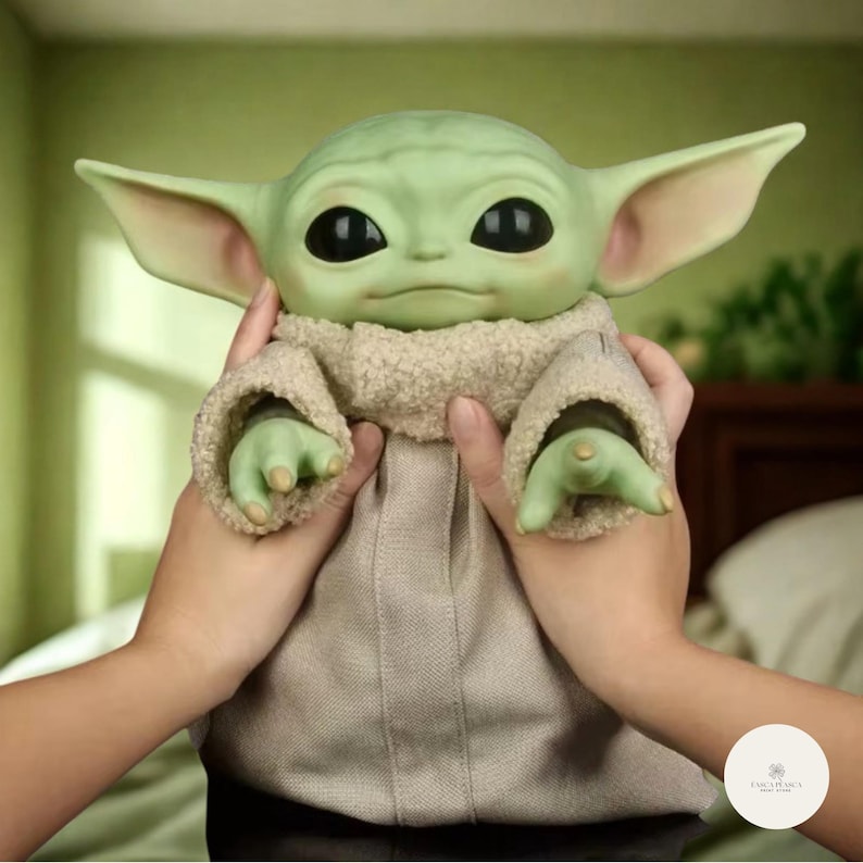 Puede incluir: Mu&ntilde;eco de piel verde con grandes orejas y grandes ojos negros, vestido con una t&uacute;nica beige. El mu&ntilde;eco est&aacute; sujeto por dos manos. El personaje es de una popular franquicia de ciencia ficci&oacute;n.