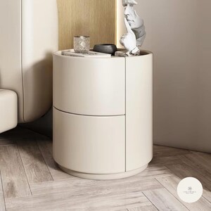 Può includere: Comodino moderno color crema con due cassetti. Il comodino ha una forma arrotondata e una piccola scultura decorativa sulla parte superiore. Un bicchierino e una ciotola scura sono anch'essi sopra. Il tutto è posto su un pavimento in legno chiaro.
