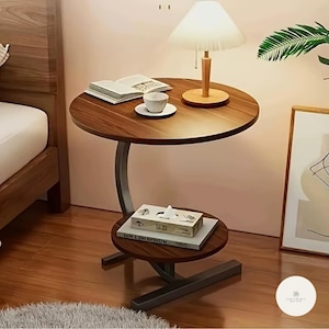 Può includere: Un tavolino in legno marrone scuro a due livelli con un supporto in metallo curvo. Il livello superiore contiene una lampada, un libro e una tazza e un piattino. Il livello inferiore ha una scatola di fazzoletti e libri.