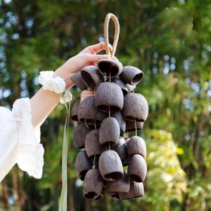 May include: A cluster of dark brown, bell-shaped ornaments, possibly made from natural materials, hangs from a woven rope. The bells are arranged in a cascading pattern, with a tag visible. The background is a blurred green, suggesting an outdoor setting.