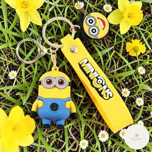 May include: A yellow Minions keychain with a cartoon character charm, a yellow strap with the word "Minions", and a round charm with a smiling face. The keychain is set against a background of green grass, yellow flowers, and small white flowers.