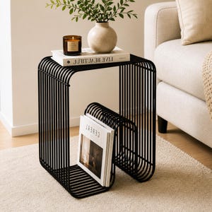 Puede incluir: Una mesa auxiliar de metal negro con un diseño de barras verticales. La mesa tiene una estantería con libros, una vela y un jarrón con vegetación. Un revistero contiene revistas. La mesa está sobre una alfombra de color claro.