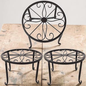 Puede incluir: Tres soportes de plantas de metal negro. Los soportes tienen una parte superior circular con un diseño calado decorativo y patas curvas. Están sobre una superficie de madera.