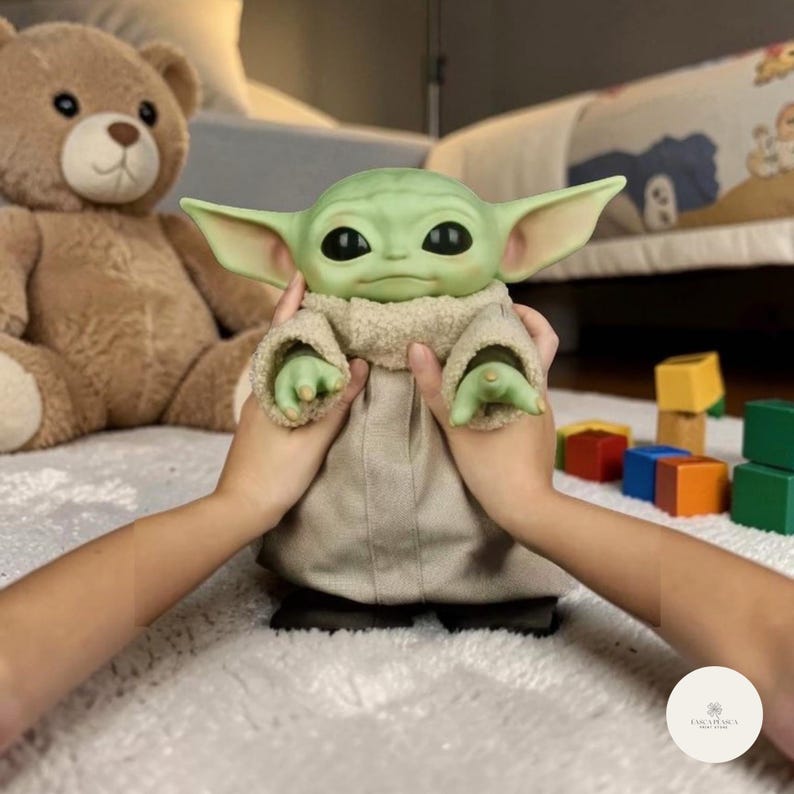 Puede incluir: Mu&ntilde;eco de personaje de piel verde con orejas grandes, con una t&uacute;nica beige y un cuello difuso. El mu&ntilde;eco est&aacute; sostenido por dos manos. Un oso de peluche marr&oacute;n y bloques de juguete coloridos est&aacute;n en el fondo.