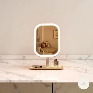 May include: A lighted vanity mirror with a rectangular shape and rounded corners. The mirror has a white frame with a built-in LED light. A small tray at the base holds a lipstick and a perfume bottle. The mirror is set on a white marble surface.