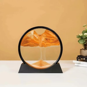 May include: A decorative sand art piece featuring a circular glass frame with a black base. The sand inside forms a mountain-like landscape in shades of orange and tan. The piece is designed to be flipped to create new patterns.