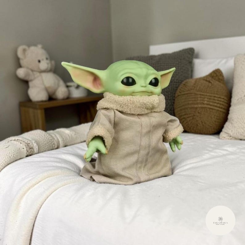 Puede incluir: Una mu&ntilde;eca de piel verde con orejas grandes, vestida con una t&uacute;nica color canela con cuello difuso, est&aacute; sentada sobre una colcha blanca. La mu&ntilde;eca tiene ojos negros grandes y manos verdes peque&ntilde;as. Un osito de peluche y cojines decorativos est&aacute;n en el fondo.
