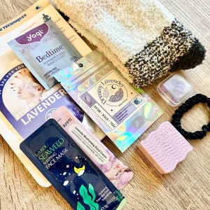 May include: A selection of relaxation and self-care products, including fluffy socks, a face mask, and lavender-infused items. The arrangement includes a tea sachet and is presented on a wooden surface, evoking a spa-like atmosphere.