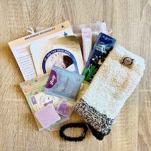 May include: A selection of relaxation items, including a face mask, eye mask, tea, fluffy socks, and a hair tie. The image also shows a small bag of lavender-scented items, a nail separator, and a leaflet on breathing techniques.