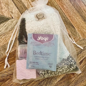 May include: A gift set in a sheer white bag, containing a pair of fluffy socks, a tea bag, and two small, rectangular items. The tea bag is labelled "Yogi Bedtime" and the socks are light blue and white.