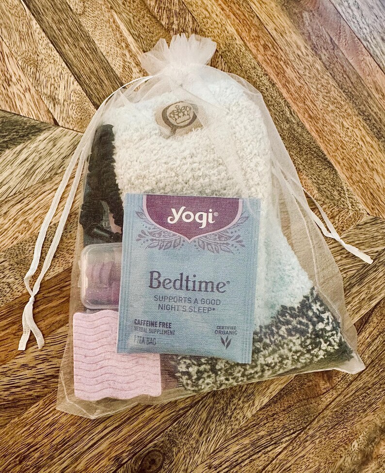 May include: A gift set in a sheer white bag, containing a pair of fluffy socks, a tea bag, and two small, rectangular items. The tea bag is labelled "Yogi Bedtime" and the socks are light blue and white.