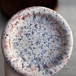 May include: A handmade ceramic plate with a speckled design. The plate has a cream base with dark blue, red, and brown speckles. The rim has a reddish-brown hue. The plate is round and glazed.