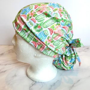 May include: A scrub cap featuring a white background with a repeating pattern of green pickles wearing colorful scarves and Santa hats, interspersed with red berries, green pine needles, and pink poinsettia flowers, tied at the back.