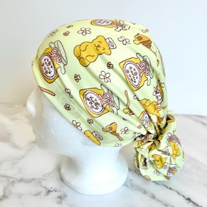 May include: A light yellow scrub cap featuring a repeating pattern of honey jars, yellow teddy bears, small bees, and white flowers. The fabric head covering has a gathered back section with a tie closure, displaying a consistent print throughout.