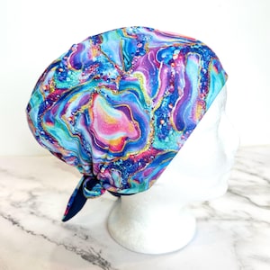 May include: A colorful scrub cap with a vibrant, swirling pattern in shades of blue, purple, pink, and gold. The cap is tied at the back and is displayed on a white mannequin head. The fabric has a marbled effect.