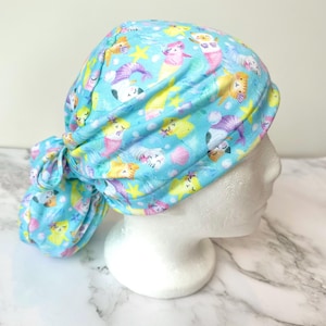 May include: A light blue scrub cap featuring a repeating pattern of cartoon cats with mermaid tails, yellow starfish, and pink seashells. The fabric head covering has a gathered back section with a tie closure, designed for a secure fit.