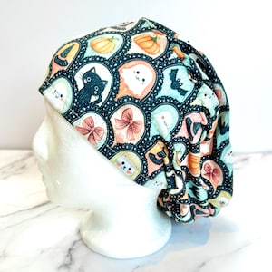 May include: A scrub cap featuring a dark teal background with a repeating pattern of Halloween-themed icons including black cats, orange pumpkins, white ghosts, bats, and pink bows, all enclosed within scalloped, star-dusted circular frames.