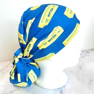 May include: A blue scrub cap featuring a repeating pattern of yellow butter sticks with the word "BUTTER" printed on them. The cap is tied at the back and is displayed on a white mannequin head.