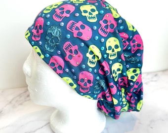 May include: A dark blue scrub cap featuring a repeating pattern of pink and yellow skulls, with clear gem embellishments. The cap is displayed on a white mannequin head.
