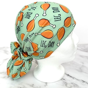 May include: A light green scrub cap with a repeating pattern of orange drumsticks and the words "LEG DAY" in black. The cap is tied in a bun at the back and is displayed on a white mannequin head.