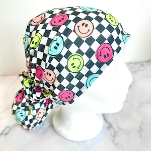 May include: A black and white checkered scrub cap with colorful smiley faces. The cap features a ponytail holder in the back and is designed to be worn during medical procedures or in a healthcare setting.