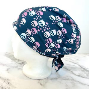 May include: A dark teal scrub cap featuring a repeating pattern of white and light pink skulls, each adorned with a small bow, interspersed with tiny pink stars. The cap has a tie-back closure at the nape of the neck and a rounded crown.