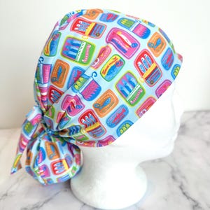 May include: A light blue surgical scrub cap featuring a repeating pattern of colorful sardine cans. The cans are in various colors, including pink, orange, yellow, and green, with the word "Sardines" printed on them. The cap is tied at the back.