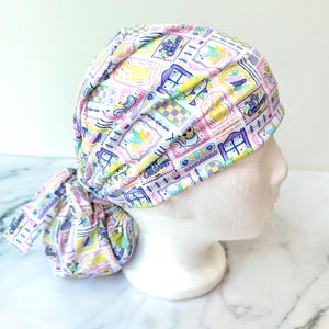 May include: A white scrub cap with a colorful pattern of framed illustrations, including hearts, houses, and text. The cap is tied in a knot at the back. The color palette includes pastel shades of pink, yellow, purple, and blue.