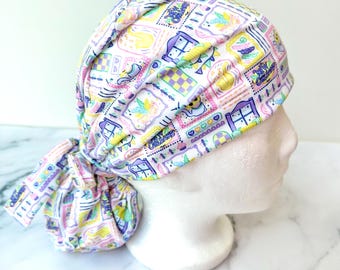 May include: A white scrub cap with a colorful pattern of framed illustrations, including hearts, houses, and text. The cap is tied in a knot at the back. The color palette includes pastel shades of pink, yellow, purple, and blue.