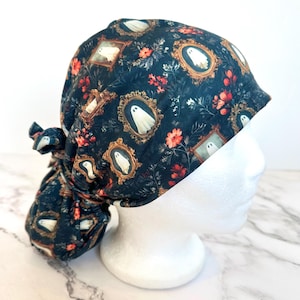 May include: A dark teal scrub cap featuring a repeating pattern of white ghosts encased in ornate gold-colored picture frames, interspersed with small red and orange floral accents. The cap includes a tie-back closure at the rear for adjustment.