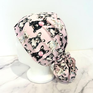 May include: A pink scrub cap featuring a repeating pattern of French bulldogs in various poses, some with bubble gum. The cap is tied at the back and is designed to fit over the hair. The fabric is a light pink color.