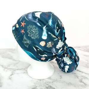 May include: A dark teal scrub cap featuring an ocean-themed pattern. The design includes illustrations of orcas, stingrays, starfish, coral, and seashells. The cap is tied at the back, and is displayed on a white mannequin head.